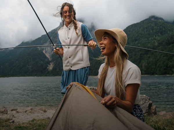 Zwei lachende Frauen in einer Berglandschaft bauen ein Zelt auf